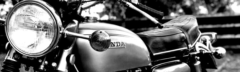 Black and white banner image of the iconic 1969 Honda CB750 motorcycle. The round headlamp, part of the handlebars, tank, and seat are visible.